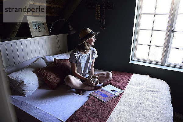 Lächelnde junge Frau sitzt auf dem Bett mit Kaffeetasse und Zeitschrift
