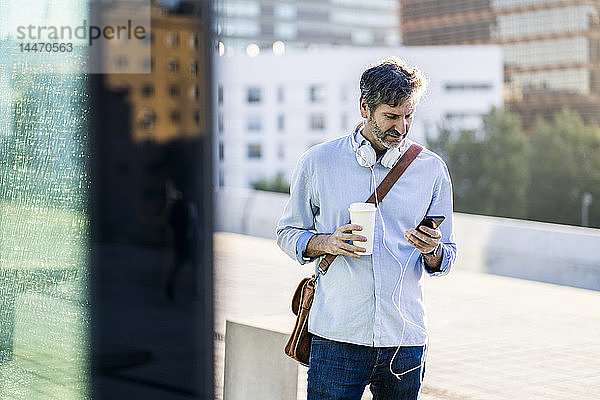 Älterer Mann mit Kaffee zum Mitnehmen und Kopfhörern  der sein Handy in der Stadt benutzt