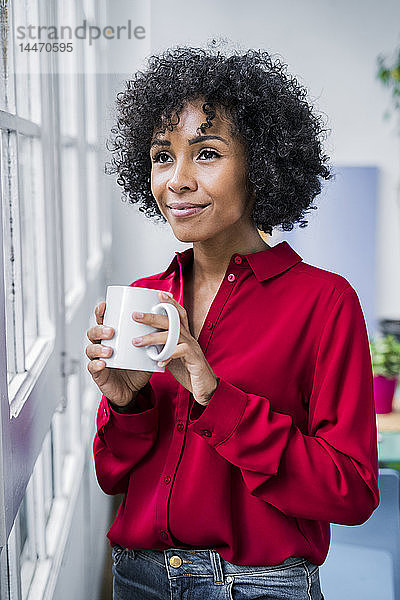 Lächelnde Frau mit Tasse Kaffee  die zu Hause aus dem Fenster schaut