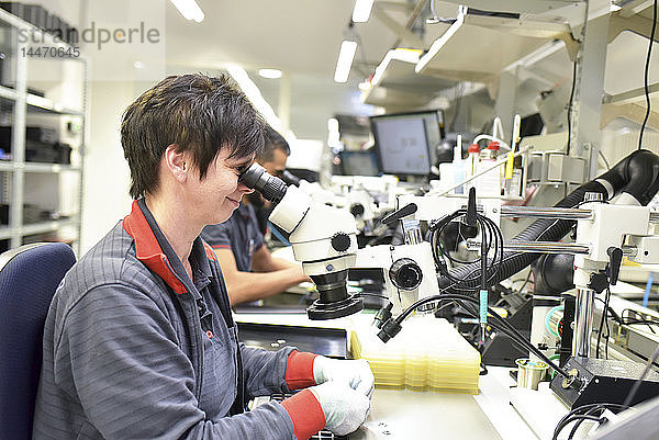 Frau  die ein Mikroskop für die Qualitätskontrolle bei der Herstellung von Leiterplatten für die Elektronikindustrie benutzt