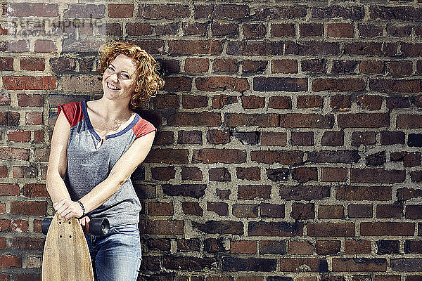 Lächelnde junge Frau mit Longboard an Ziegelmauer stehend