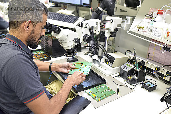 Mann  der ein Mikroskop für die Qualitätskontrolle bei der Herstellung von Leiterplatten für die Elektronikindustrie benutzt
