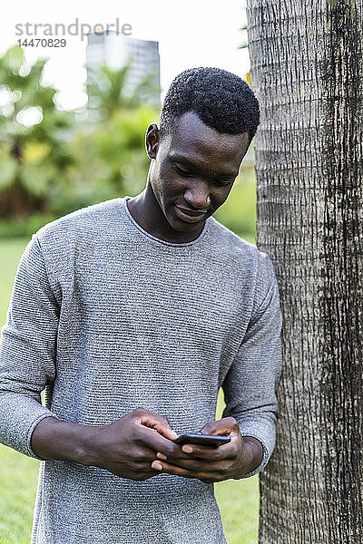 Junger Mann lehnt sich in einem Park an einen Baumstamm und benutzt ein Smartphone