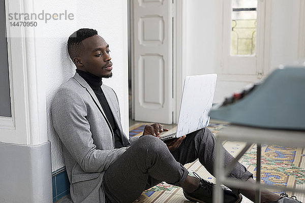 Porträt eines jungen Geschäftsmannes  der im Büro auf dem Boden sitzt und am Laptop arbeitet
