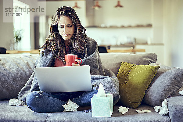 Eine kranke Frau sitzt auf einem mit einer Decke bedeckten Sofa mit einer Tasse Tee und einem Laptop