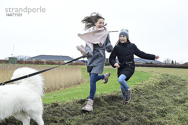 Zwei Mädchen rennen mit Hund auf einer Wiese und amüsieren sich