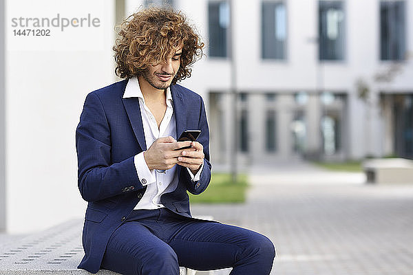 Lächelnder junger Geschäftsmann mit lockigem Haar im blauen Anzug sitzt auf Bank im Freien und schaut auf Handy