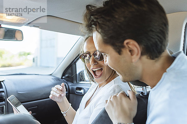 Glückliches Paar im Auto mit Mann auf dem Rücksitz und Frau mit Handy auf dem Beifahrersitz
