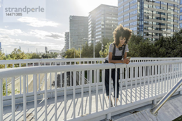 Junge Frau steht auf einer Brücke  benutzt ein Smartphone und hat Kopfhörer um den Hals
