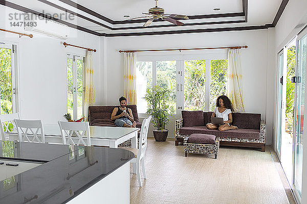 Thailand  Ehepaar sitzt in geräumigem Wohnzimmer am Ferienort und benutzt elektronische Geräte