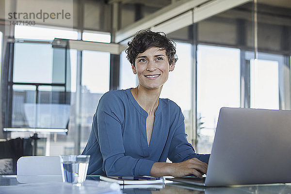 Lächelnde Geschäftsfrau sitzt mit Laptop am Schreibtisch im Büro