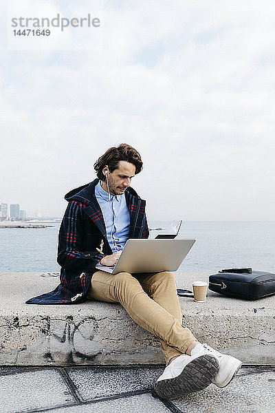 Spanien  Barcelona  Mann sitzt am Meer und arbeitet mit Laptop und Notizbuch