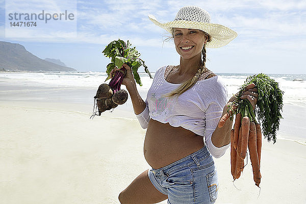 Porträt einer lächelnden schwangeren Frau am Strand mit Karotten und Rote Beete