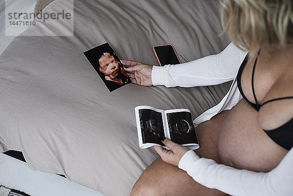 Schwangere Frau betrachtet Ultraschallbilder zu Hause im Sitzen auf dem Bett