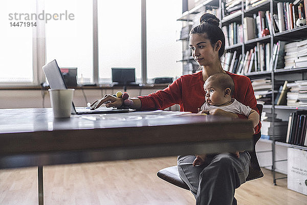 Berufstätige Mutter mit Baby auf dem Schoß  im Büro sitzend  mit Laptop
