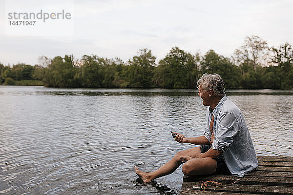 Glücklicher älterer Mann sitzt mit Handy auf einem Steg an einem See