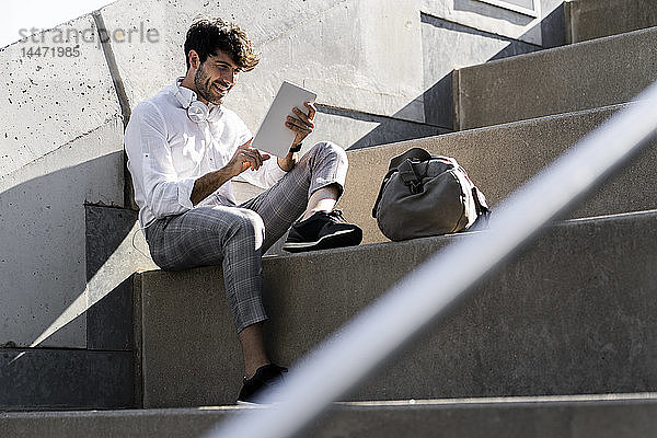 Lächelnder junger Mann sitzt mit Tablette auf einer Treppe im Freien