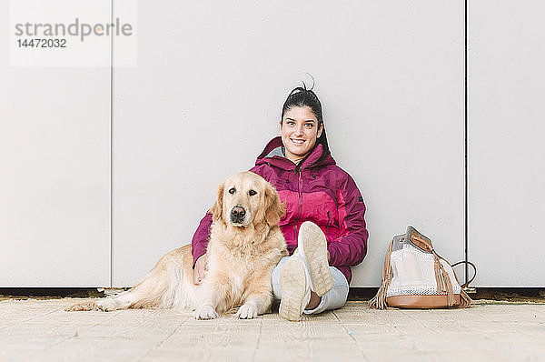 Porträt einer lächelnden jungen Frau mit ihrem Golden-Retriever-Hund an einer Wand sitzend