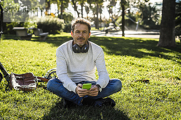 zufälliger Geschäftsmann  der auf einer Wiese in einem Park sitzt und ein Smartphone benutzt