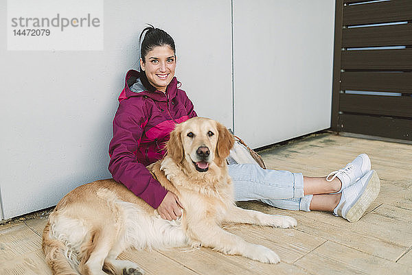 Porträt einer lächelnden jungen Frau mit ihrem Golden-Retriever-Hund an einer Wand sitzend