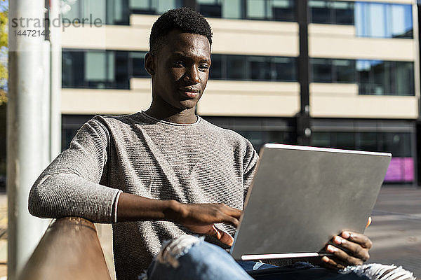 Junger Mann sitzt auf einer Bank in der Stadt und benutzt einen Laptop