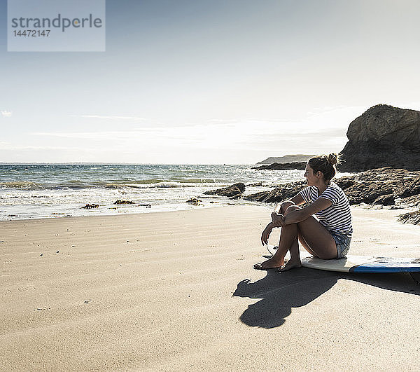 Junge Frau sitzt auf ihrem Surfbrett am Strand und schaut auf das Meer