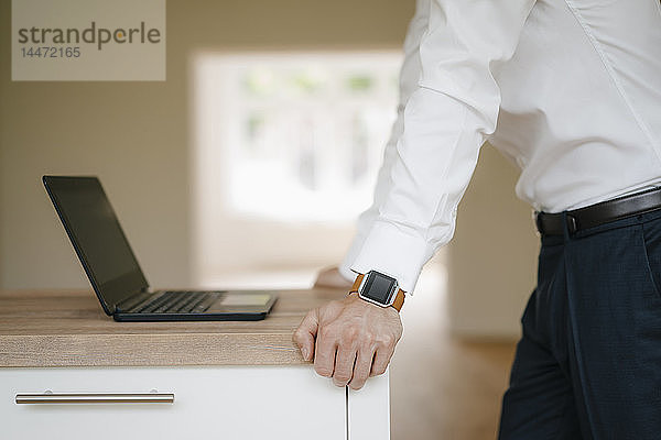 Geschäftsmann lehnt sich auf Kitchen-Oberfläche  benutzt Laptop  trägt smarte Uhr