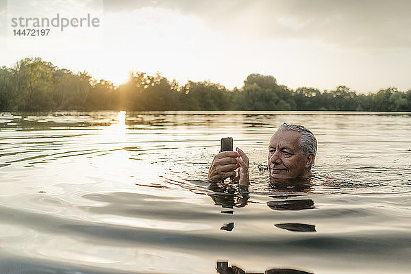 Älterer Mann schwimmt bei Sonnenuntergang mit seinem Handy in einem See