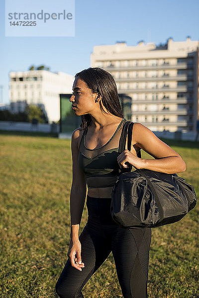 Sportliche junge Frau mit Tasche steht auf der Wiese der Stadt