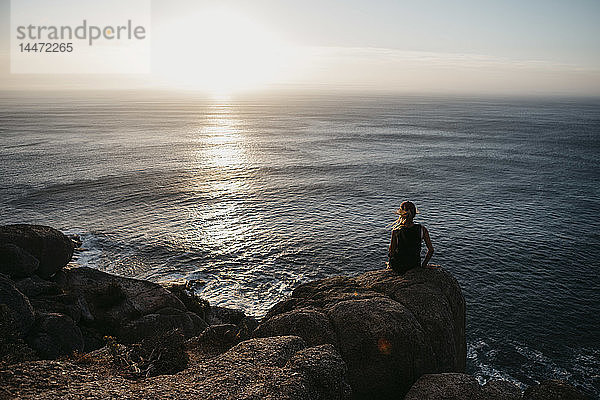 Südafrika  Westkap  Frau sitzt auf einem Felsen und betrachtet den Sonnenuntergang  vom Chapman's Peak Drive aus gesehen