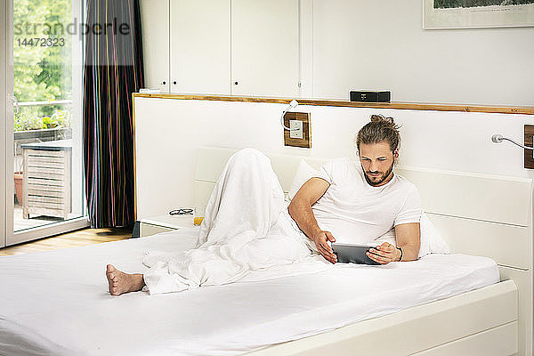 Junger Mann liegt im Bett und benutzt morgens ein digitales Tablett