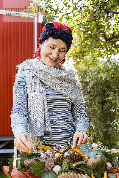 Porträt einer lächelnden älteren Frau mit rot gefärbten Haaren beim Basteln an der Herbstdekoration im Garten