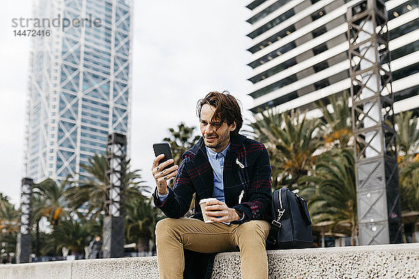 Spanien  Barcelona  lächelnder Mann sitzt in der Stadt mit Kaffee zum Mitnehmen und Mobiltelefon