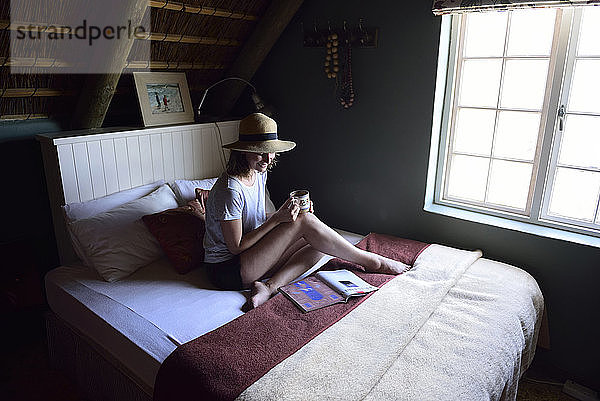 Lächelnde junge Frau sitzt auf dem Bett mit Kaffeetasse und Zeitschrift