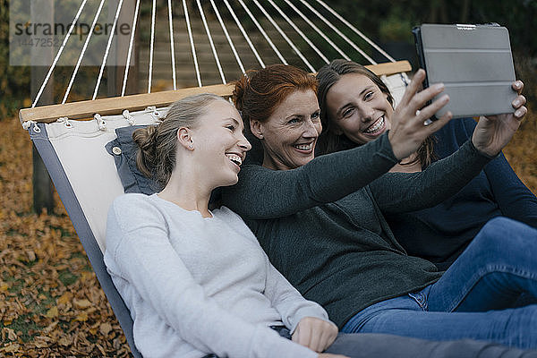 Glückliche Mutter mit zwei Teenager-Mädchen  die im Herbst in der Hängematte im Garten liegen und Tabletten benutzen