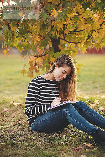 Junge Frau mit Notizbuch sitzt im Herbst auf einer Wiese in einem Park und macht Notizen