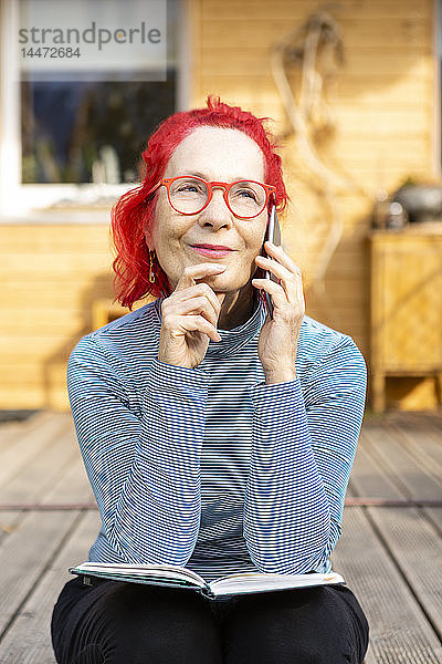 Porträt einer lächelnden älteren Frau mit rot gefärbten Haaren am Telefon  die auf der Terrasse vor ihrem Haus sitzt