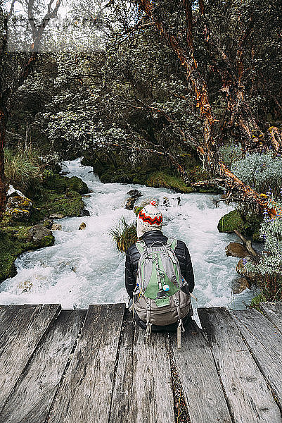 Peru  Huaraz  Mann mit Wollmütze und Rucksack auf Holzbrücke an einem Fluss sitzend