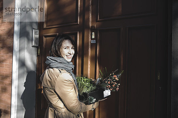 Porträt einer lachenden Frau mit Pappkarton mit Einkäufen vor dem Hauseingang stehend