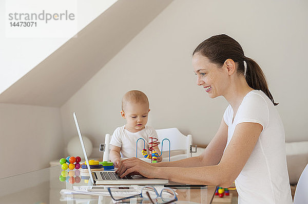 Lächelnde Mutter benutzt Laptop und kleine Tochter spielt zu Hause am Tisch