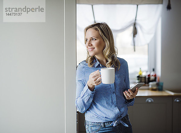 Lächelnde Frau hält zu Hause eine Tasse Kaffee und ein Smartphone