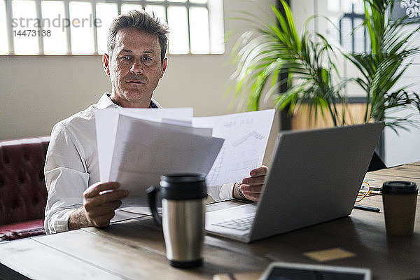 Fokussierter Geschäftsmann  der einen Laptop benutzt und Dokumente am Schreibtisch durchsieht