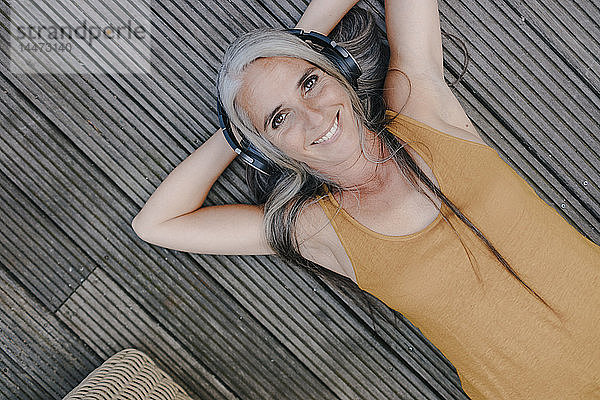 Porträt einer glücklichen Frau  die auf der Terrasse liegt und mit Kopfhörern Musik hört