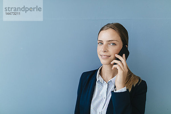 Porträt einer jungen Geschäftsfrau vor blauem Hintergrund  die auf ihrem Smartphone spricht