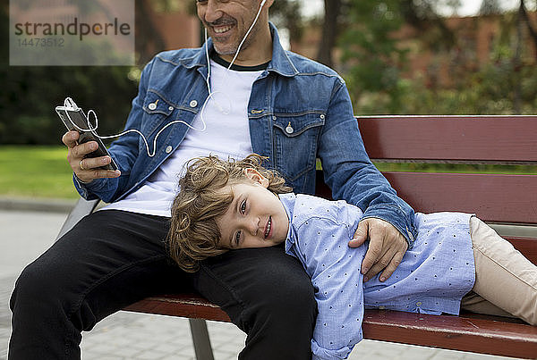 Porträt eines lächelnden Jungen mit Vater  der mit Handy und Ohrstöpseln auf einer Bank sitzt