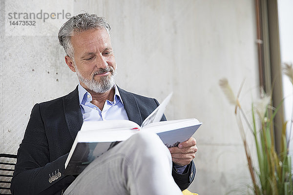 Lächelnder Geschäftsmann sitzt an Betonwand und liest Buch