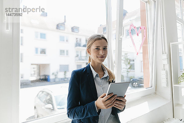 Junge Geschäftsfrau steht am Fenster und benutzt ein digitales Tablet