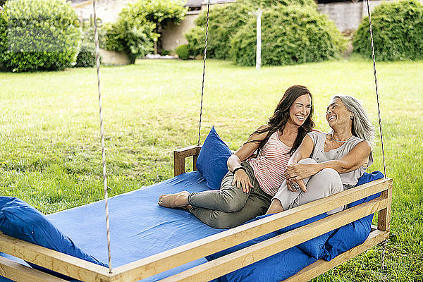 Zwei glückliche Frauen entspannen sich auf einem hängenden Bett im Garten