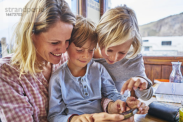 Glückliche Mutter mit zwei Söhnen  die in einem Café am Tisch sitzen und mit dem Handy spielen