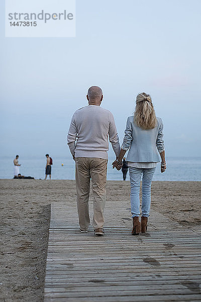 Spanien  Barcelona  Rückansicht eines älteren Ehepaars  das in der Abenddämmerung Hand in Hand am Strand spazieren geht